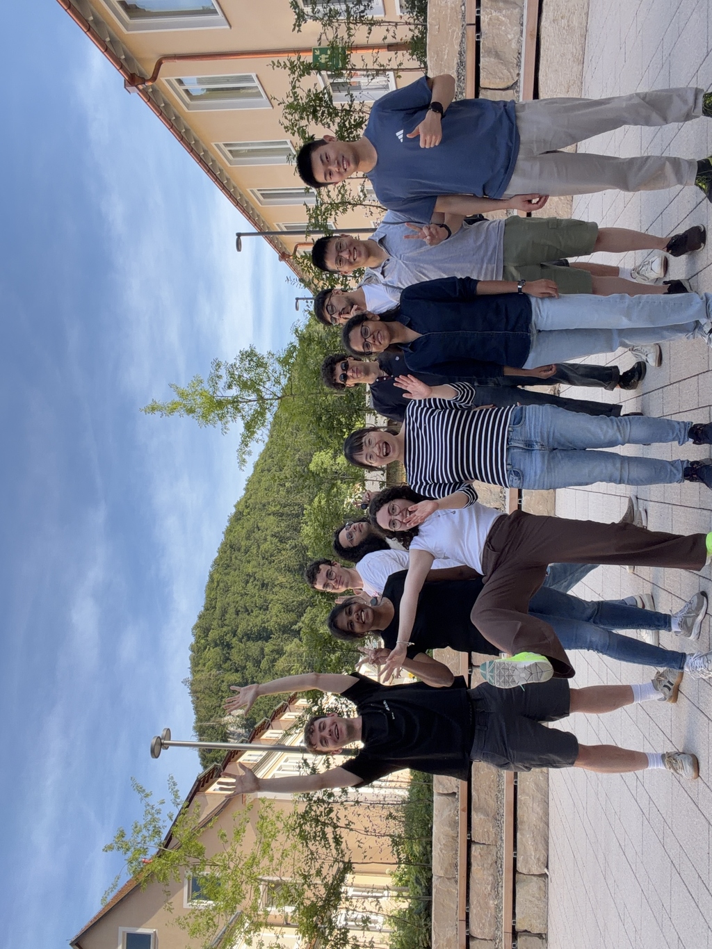 Group of young adults posing for a photo outdoors near a building and a forested hill.