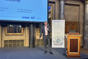 A man giving a presentation at a VDE VDI GMM conference next to a podium and a banner.